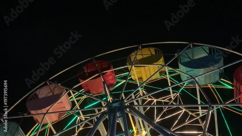 Close-up shot of a colorful Ferris wheel at night with vibrant green, yellow, and orange lights against a dark background