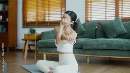 Happy young Asian woman practicing yoga and shoulder stretching at home sitting on floor in living room
