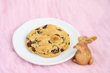 Celebrate Easter holiday with a large gourmet chocolate chip cookie with gold decorative bunny looking at the treat on a white plate, on soft pink fabric background
