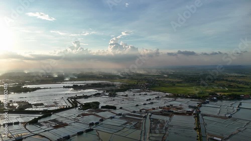 Aerial View of Traditional Salt Evaporation Ponds Bordering Green Rice Fields and Village Settlement under Cloudy Sky