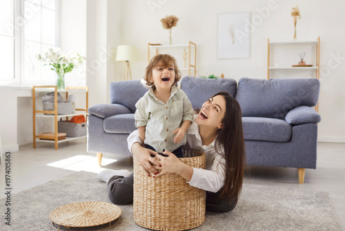 Happy mother and child play at home, bonding. A playful toddler kid laughs in a wicker basket as woman parent smiles nearby, sharing love and happiness. Warm family leisure and joy together.