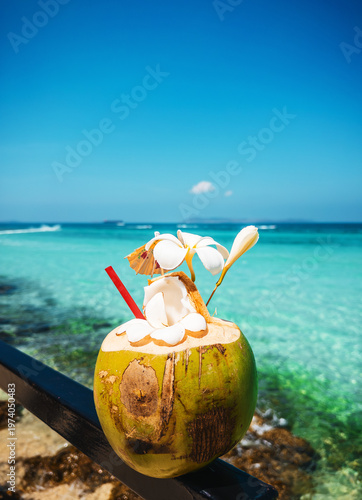 Fresh coconut drink with straw and tropical flowers on seaside background. Exotic vacation mood with turquoise water, sunny beach. Concept of summer rest, travel and relaxation. Early travel booking.