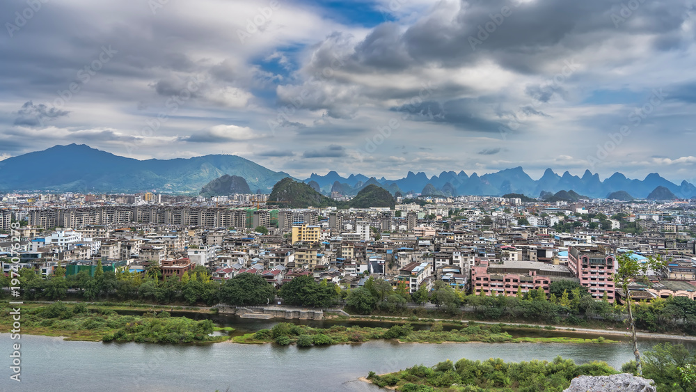 Obraz premium Panorama of a modern city. View from the height. There are many multi-storey buildings, an embankment. Green vegetation on the river bank. Picturesque mountains on the horizon. Blue sky, clouds.China