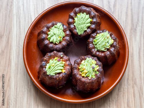Close-up of gourmet mini chocolate canelé-style desserts topped with green pistachio cream and crushed nuts, served on a rustic brown ceramic plate. Artistic pastry presentation