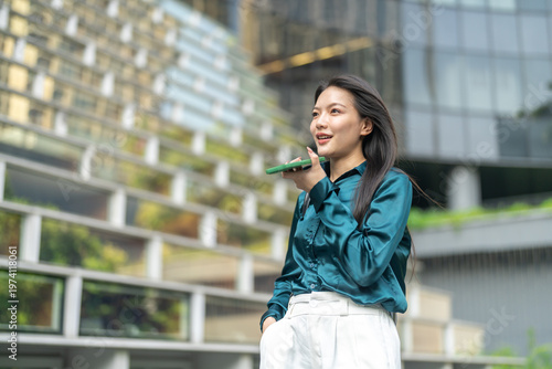 Smiling asian business woman using AI voice assistant on smartphone outdoors, Smart agent technology, Female corporate employee recording audio message or speaking to artificial intelligence app