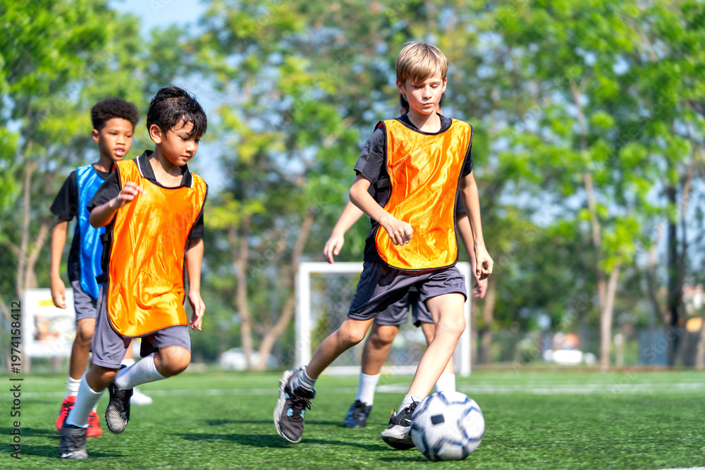 Fototapeta premium A boy with blond hair try to dribble the ball to the other side away from the competitor team during practice of soccer in the field with day light.