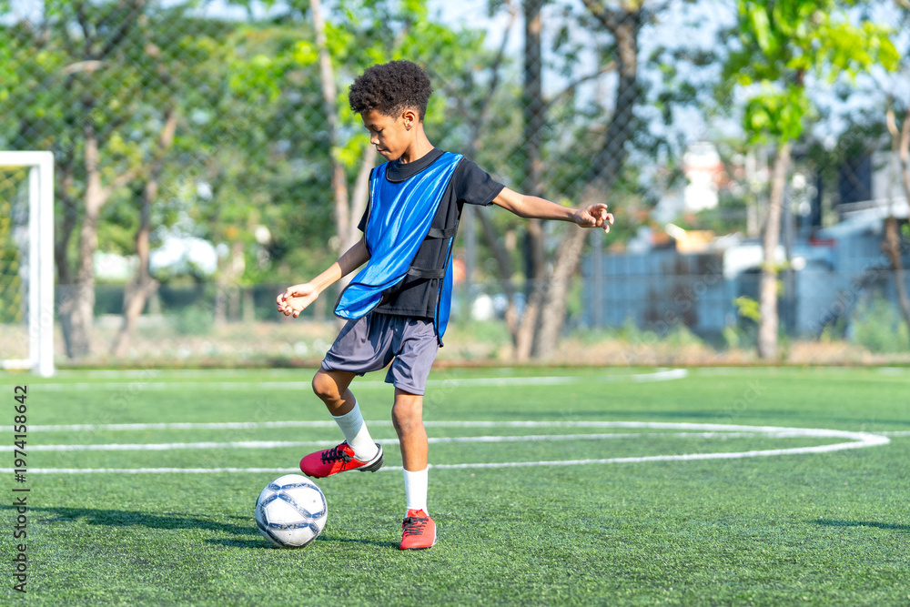 Obraz premium A little african-american boy is dribbling the football in the field to play soccer alone during day light to practice and prepare for competitiion.