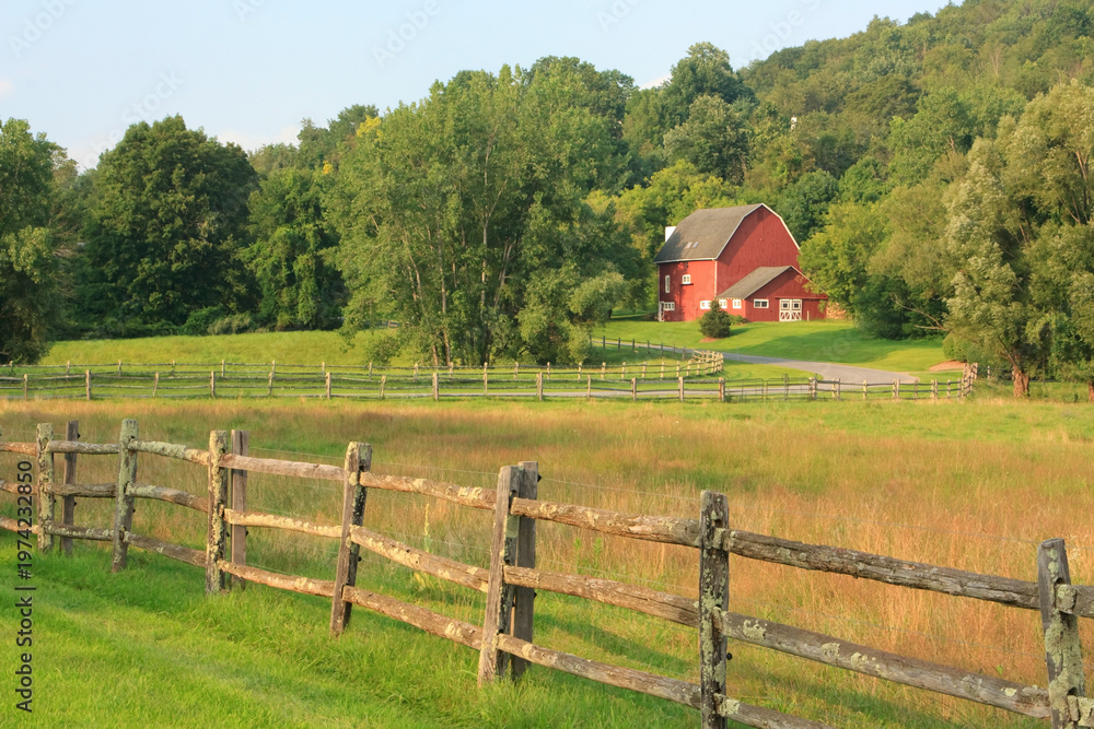 Fototapeta premium Rural summer landscape near Kent in Connecticut with a rail fence in the front and a red barn in the distance