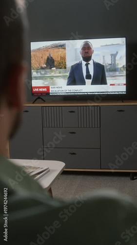 Vertical over-shoulder rack focus shot of male televiewer with remote control turning on TV news broadcast on overseas military invasion or international war conflict, soldiers running into battle