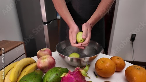 A man in an apron stands in front of a table with fruit, washing green apples in a metal bowl filled with water before cooking. Close up, front view.