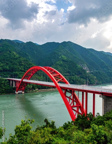 A vibrant red arch bridge gracefully spans a wide river, nestled amidst lush green mountains under a partly cloudy sky, showcasing impressive engineering and scenic beauty.