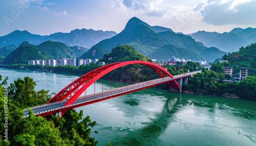 A vibrant red arch bridge gracefully spans a wide river, nestled amidst lush green mountains and a modern cityscape under a partly cloudy sky.