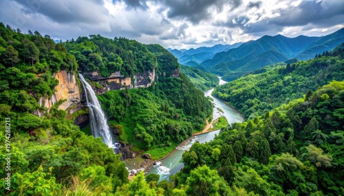 A breathtaking panoramic view showcases a cascading waterfall plunging into a winding river, surrounded by lush green mountains under a dramatic cloudy sky.