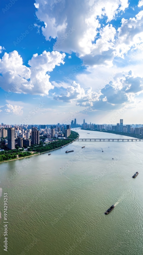 Obraz premium An aerial view showcases a sprawling cityscape nestled along a wide river, framed by a vibrant blue sky dotted with fluffy white clouds and a modern bridge connecting the urban landscape.
