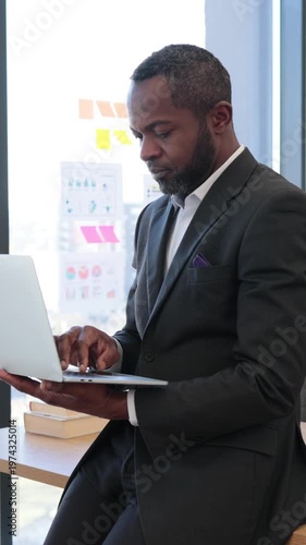 A focused African American businessman in a suit works diligently on his laptop while sitting near a window with charts on the wall