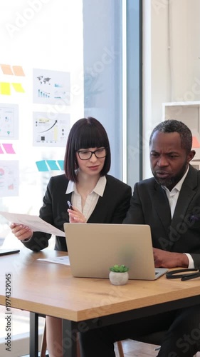 A woman and an African American man collaborate on a project using a laptop and documents in a modern office setting