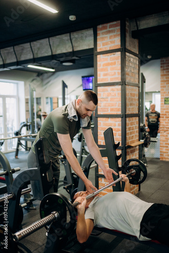A man lies on a bench pressing weights while another man assists him in a gym. The atmosphere is busy with people working out and equipment around.