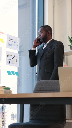 An African American businessman in a suit talks on his phone while looking at charts and graphs on a glass wall