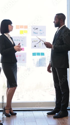 A woman and an African American man in business attire discuss charts and graphs on a glass board during a meeting