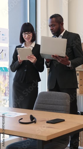 A woman and an African American man in business attire collaborate using a tablet and laptop in a modern office setting
