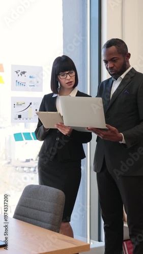 A woman and an African American man in suits collaborate on a project using a laptop and tablet in a modern office setting