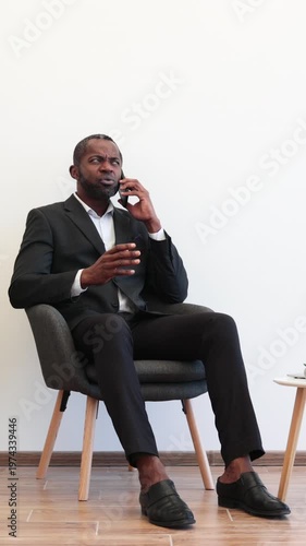 An African American man in a suit sits in a chair while talking on his phone with a perplexed expression, gesturing with his hand