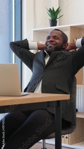 An African American man in a suit leans back in his office chair with hands behind his head, looking up with a satisfied expression