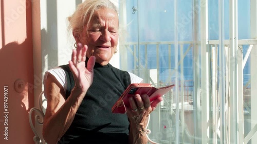Senior woman waving during a video call on her smartphone. Concept of family connection, digital literacy for seniors, and remote communication with grandchildren via mobile technology