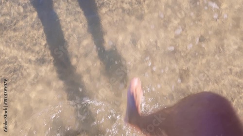 Close top view of male feet walking on sand with waves washing over. Bright sunlight and motion evoke freedom, calmness and a peaceful coastal vibe.
