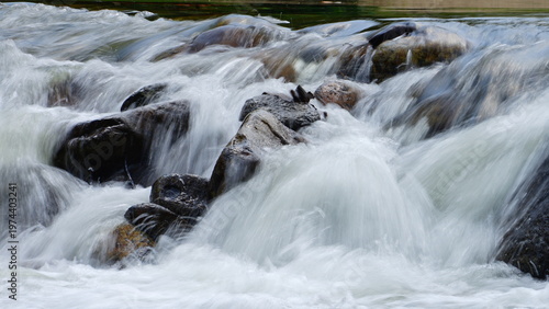 Close-up view of fast-flowing water cascading over smooth rocks in a natural stream. The motion blur creates a soft, silky effect, highlighting the dynamic movement and energy of the rushing current. 