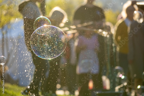 Close-up of a sparkling soap bubble in golden sunset light, with blurred people and splashing droplets in the background, creating a dreamy festive outdoor scene.