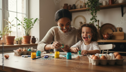Happy african american mother and daughter painting easter eggs in kitchen with face paint