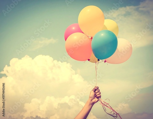 A person's arm holds a bunch of colorful balloons against a cloudy, bright, vintage-filtered sky