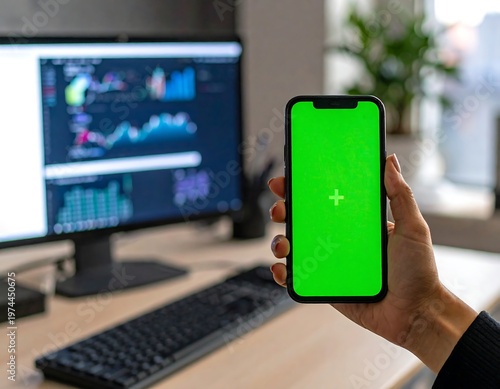 A person's hand holds a phone with a green screen, in front of a computer monitor displaying charts and graphs