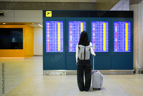 A young woman with backpack and suitcase stands in front of a digital destination board at airport. with his back to the camera. A tourist studying the arrival of flights on an electronic scoreboard