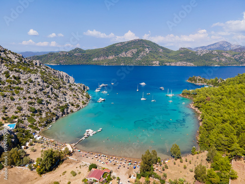 Photos Sig Liman Bay in Selimiye, Marmaris, Mugla, Türkiye, features turquoise waters, anchored boats, and a lively beach surrounded by forested hills and Mediterranean scenery