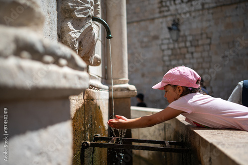 Little girl drinks water from The Large Onofrios Fountain in old town of Dubrovnik, Croatia. Thirsty girl drinks water from a fountain. 