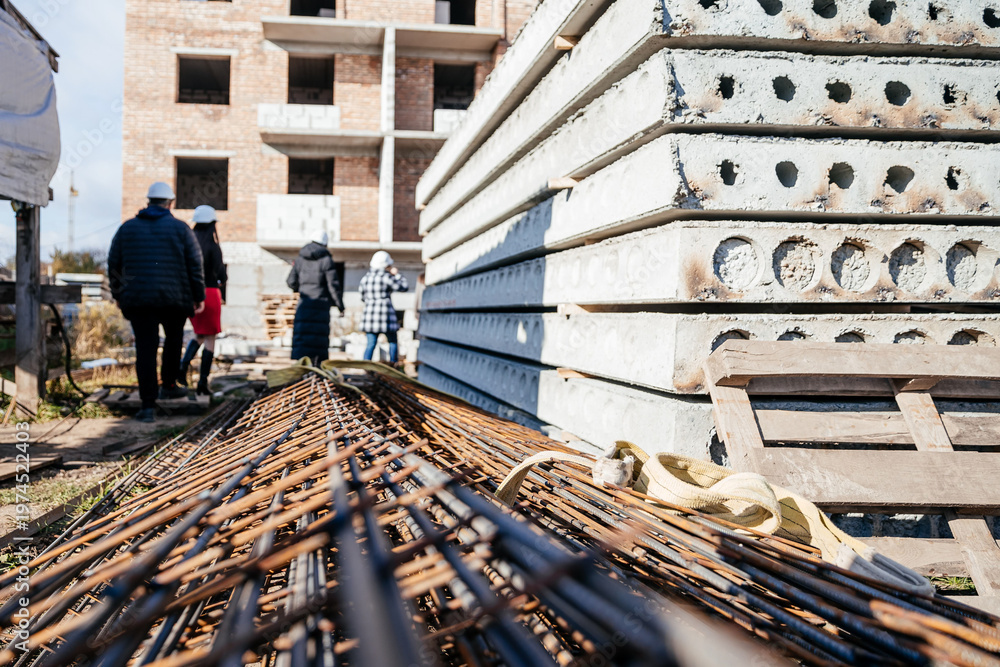 Naklejka premium Workers walk past stacks of concrete blocks and steel rods at a new building site under bright sunlight