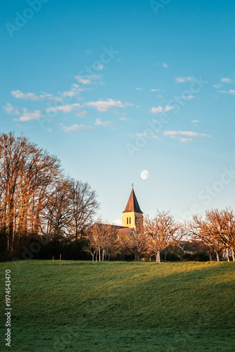 Three quarter moon over the spire of the village church at Nabirat in the Dordogne region of France lit by the early morning sun