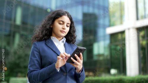 Businesswoman standing outside an office building while using her smartphone. Female employee looks serious and focused on her mobile phone, checking information in an urban environment, close up.