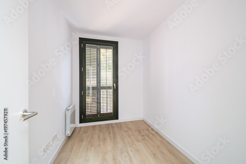 Empty room with white walls and wooden floor. A large window with black frames allows natural light to enter. Minimalist design with no furniture.