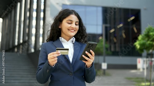 Smiling businesswoman shops online using a credit card and smartphone while standing near office building. Female employee enjoys convenient digital purchase experience in urban environment, close up.