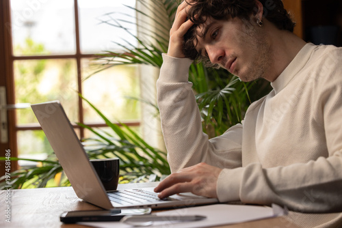 Jeune homme en situation de stress face à son ordinateur, illustrant la pression du télétravail et la fatigue mentale dans un environnement domestique lumineux.