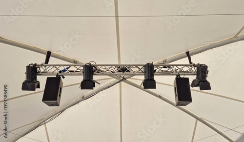 A row of black professional spotlights and audio speakers hanging from a metal truss system against the white canvas background of a large event tent