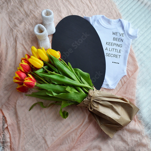 Baby announcement with white bodysuit, knit booties and tulip bouquet. Pregnancy disclosure message on blackboard with floral decoration. Concept of impending birth and new life event.