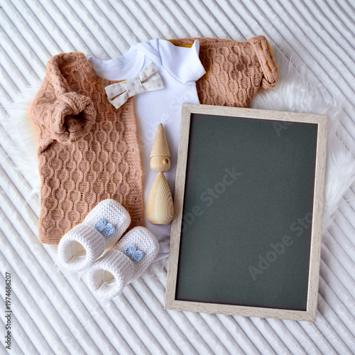 Baby clothes set with chalkboard for announcement. Knitted sweater, bodysuit, bow tie and booties with wooden toy on fur background. Nursery decoration and newborn greeting card concept.