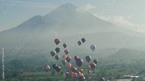 A stunning aerial scene of multiple colorful hot air balloons drifting peacefully above a lush countryside with a majestic mountain backdrop.