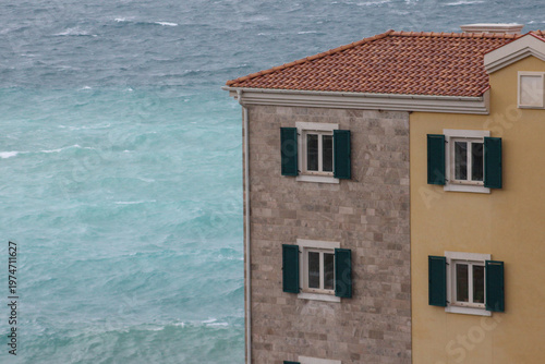 Foto Close view of a Mediterranean coastal house with stone and yellow walls, green shutters and tiled roof, set against a vibrant turquoise sea with waves on a breezy day