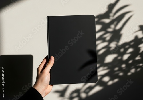 A hand holding a blank black notebook cover against a wall with artistic palm leaf shadows cast