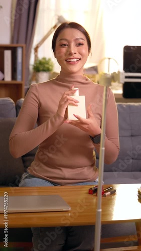 Confident asian beauty influencer woman demonstrating skincare cosmetic product to camera feeling enthusiastic recording vertical live broadcast sitting comfortably on cozy living room sofa